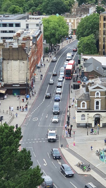An aerial view of a street in Bermondsey, showing a line of parked vehicles and moving cars on a two-lane road, with trees lining both sides of the street. On the left, there's a mix of brick and modern commercial buildings, some with signage, and pavement areas where pedestrians are walking. On the right, there are residential and historic-style buildings with small front yards and bicycles parked on the sidewalk. The scene depicts typical urban activity during daytime, with a few people walking and cycling, and the road appears to be part of a home relocation or moving process managed by a local removals service such as Man and Van Bermondsey. The environment is well-lit with natural daylight, and the overall setting indicates an active, residential neighbourhood suitable for furniture transport and packing and moving activities.