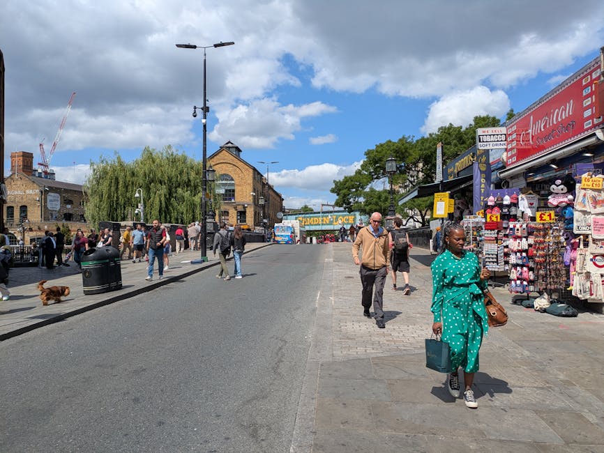 A busy street scene on Bermondsey High Street with pedestrians walking along the pavement, some carrying shopping bags, and a woman dressed in a teal polka-dot outfit holding a handbag and a shopping bag. Several other individuals are visible, including a man in a beige jacket and black pants. To the right, there is a street stall displaying various items such as hats, souvenirs, and household goods, with price tags visible. The shopfront signage includes a red sign for 'American Street Shop' and other smaller signs for local businesses. The street is lined with lampposts, and the background features a brick building with large windows, some trees, and blue sky with scattered clouds. This scene illustrates a typical urban environment where house removals and furniture transport services by Man and Van Bermondsey might operate in context of local moving logistics.