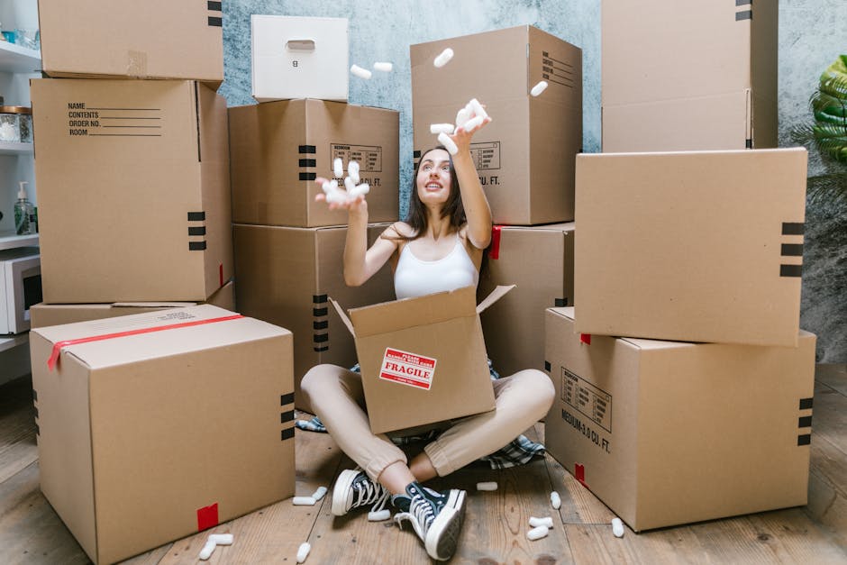 A young woman with long brown hair, dressed casually in a white sleeveless top, beige trousers, and black and white sneakers, is sitting on a wooden floor surrounded by numerous cardboard boxes used f