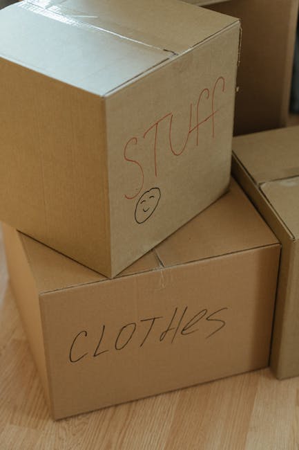 A young woman with long brown hair, dressed casually in a white sleeveless top, beige trousers, and black and white sneakers, is sitting on a wooden floor surrounded by numerous cardboard boxes used f